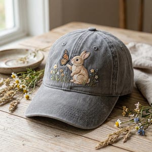 Könnte beinhalten: Graue Baseballkappe mit einem gestickten Design, das einen braunen Hasen und einen Schmetterling zeigt. Der Hase greift nach dem Schmetterling, umgeben von gestickten Blumen und Gras. Die Kappe hat einen gebogenen Schirm und einen Knopf.