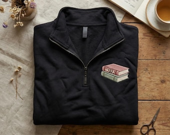 May include: Black quarter-zip sweatshirt with an embroidered design of two stacked books in red and green. The sweatshirt has a brass-colored zipper and a high collar. The background includes a wooden surface, a cup of tea, and a book.