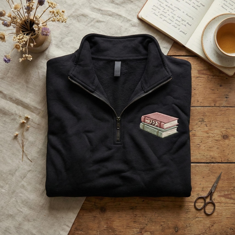 Puede incluir: Sudadera negra con cremallera de un cuarto con un dise&ntilde;o bordado de dos libros apilados en rojo y verde. La sudadera tiene una cremallera color lat&oacute;n y un cuello alto. El fondo incluye madera, t&eacute; y un libro.