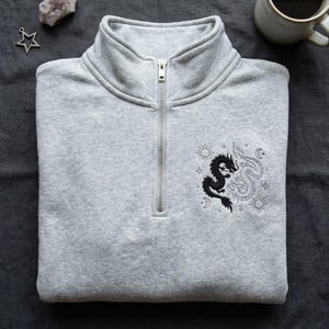 May include: A light gray quarter-zip sweatshirt with a silver zipper. The sweatshirt features an embroidered design of two dragons, one black and one white, with sun and moon details. The sweatshirt is folded neatly.