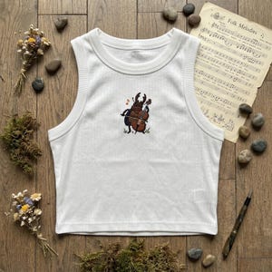 May include: A white ribbed tank top featuring a central embroidered graphic of a brown beetle playing a cello. The beetle is depicted with a small orange musical note above it. The garment has a crew neckline and a sleeveless design.