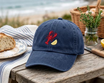 May include: Navy blue baseball cap with a red embroidered lobster and yellow lemon slice. The cap is sitting on a wooden table with a picnic setting in the background. The hat is a classic, casual style.
