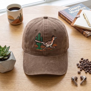 Peut inclure: Une casquette de baseball marron avec une broderie représentant un grand géocoucou à côté d'un cactus vert. La casquette présente une calotte structurée, une visière courbée et des œillets cousus sur les panneaux supérieurs, au motif désertique.
