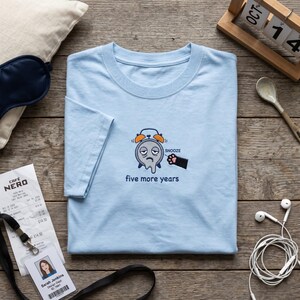 May include: A light blue short-sleeve t-shirt featuring an embroidered graphic of a melting, tired alarm clock with orange bells. A black cat paw presses the snooze button, and the text below reads five more years in blue letters.