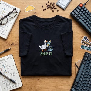 May include: A black short-sleeve t-shirt featuring an embroidered graphic of a white goose holding a small rocket ship next to an open laptop with a green checkmark on the screen. Below the graphic, the text SHIP IT is embroidered in green.