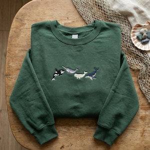 May include: A dark green sweatshirt with embroidered images of a killer whale, narwhal, and two other whales. The sweatshirt has a crew neck and long sleeves. The whales are in shades of white, grey, and blue.