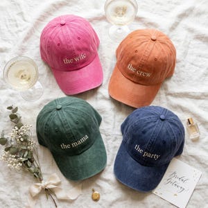May include: Four baseball caps in pink, orange, green, and blue. Each cap has white embroidered text: "the wife", "the crew", "the mama", and "the party". The caps are arranged on a white surface with champagne glasses and a perfume bottle.