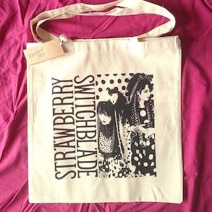 May include: A white canvas tote bag with black handles and a black and white graphic print of the band "Strawberry Switchblade". The text "Westford Mill" is printed on a small brown tag attached to the bag.