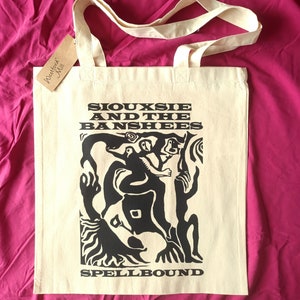May include: A white canvas tote bag with black handles and a black print of the band Siouxsie and the Banshees. The print features a stylised image of a person with a crown and a large, abstract design. The text "Spellbound" is printed below the image.