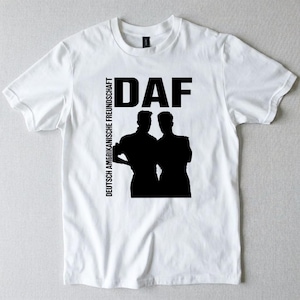 以下が含まれることがあります： 胸元に黒いグラフィックプリントが施された白い半袖Tシャツ。デザインには上部に太字でDAFの文字、垂直に書かれたDEUTSCH AMERIKANISCHE FREUNDSCHAFTの文字、そして並んで立つ2人の人物のシルエットが含まれています。