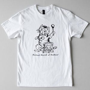 May include: White t-shirt with a black and white illustration of a cat playing a drum. The cat is shown with a drumstick and a drum. The text "Postcard Records of Scotland" is printed below the illustration, a design for music lovers.