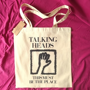 May include: A beige tote bag with black text and a graphic of a fist. The text reads "Talking Heads" and "This Must Be The Place".