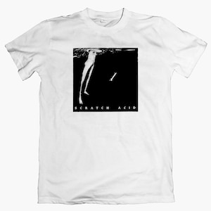 Puede incluir: Camiseta blanca con un gráfico en blanco y negro. El gráfico es una imagen rectangular con una figura y un objeto en forma de hueso sobre un fondo negro. El texto "SCRATCH ACID" está debajo.