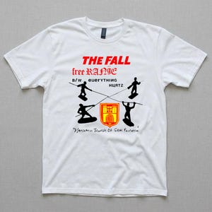 Op de afbeelding: Wit t-shirt met een grafisch ontwerp met de tekst "THE FALL" in rood, "free RANIE" in zwart, en andere tekst en afbeeldingen. Het ontwerp bevat zwarte silhouetten en een rood en geel wapenschild.