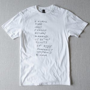 Op de afbeelding: Wit t-shirt met zwarte handgeschreven tekst. De tekst bevat woorden als "HURTZ", "TIME", "IDIOT", "MARRIED" en "B'HAM". Het shirt heeft korte mouwen en een ronde hals.