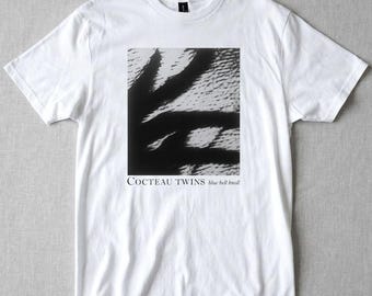 May include: White t-shirt featuring a black and white graphic print. The graphic is a textured, abstract design. The text "COCTEAU TWINS" and "blue bell knoll" are printed below the graphic.