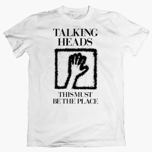 Op de afbeelding: Wit T-shirt met een zwarte afbeelding van een vuist in een vierkant met de tekst "Talking Heads" en "This Must Be The Place" eronder.
