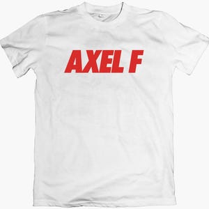 May include: White t-shirt featuring the red text "AXEL F" across the chest. The shirt has a crew neck and is likely made of cotton. The text is in a bold, clear font.