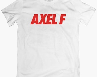 Puede incluir: Camiseta blanca con el texto rojo "AXEL F" en el pecho. La camiseta es de estilo cuello redondo clásico, probablemente de algodón. El texto es en negrita y fácil de leer.