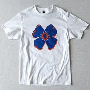 May include: White t-shirt with a blue and orange floral design. The flower graphic has a circular centre with the words "Flowered Up It's On" printed in orange. The t-shirt is made of a soft, lightweight material.