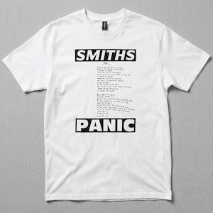 Peut inclure: T-shirt blanc avec le nom du groupe "SMITHS" et le mot "PANIC" en noir gras. Le t-shirt affiche également les paroles de la chanson "Panic" dans une police manuscrite.