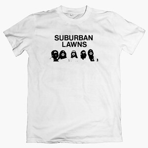 May include: White t-shirt featuring the text "SUBURBAN LAWNS" in black above a row of black, stylized figures with large eyes. A simple, graphic design.