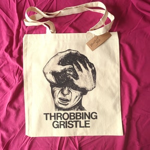 Op de afbeelding: Een beige tote bag met zwarte print. De print toont een zwart-witte illustratie van een gezicht met de hand die de ogen bedekt. De tekst "Throbbing Gristle" is onder de illustratie gedrukt. Een bruin label met de tekst "Westport Mill" is aan de tas bevestigd.