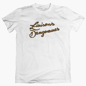 May include: A white t-shirt featuring the words "Liaisons Dangereuses" in a brown, cursive font. The text is centered on the front of the shirt. The shirt has short sleeves and a crew neck.