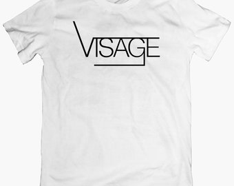 Puede incluir: Camiseta blanca con un gráfico negro que dice "VISAGE" en una fuente en negrita y sin serifas.