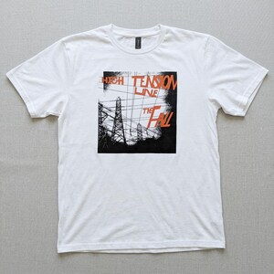 Puede incluir: Camiseta blanca con un diseño gráfico. El diseño presenta el texto "HIGH TENSION LINE" y "THE FALL" en naranja y negro. El gráfico incluye líneas eléctricas y torres.