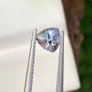 May include: A pear-shaped, pale blue gemstone held by silver tweezers. The gem's faceted surface reflects light, creating a starburst pattern. The background features blurred greenery and white surfaces.