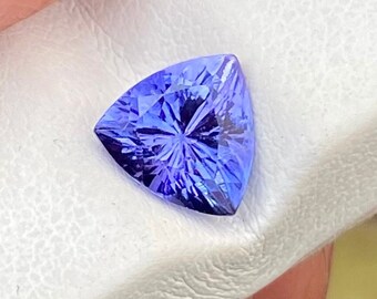 May include: A triangular cut, purple gemstone with a faceted surface. The gemstone is displayed on a white, textured surface.