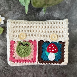 May include: A crocheted cream-colored clutch with two brown buttons. The clutch has two square panels with colorful crocheted designs. One panel features a green frog and the other a red mushroom.