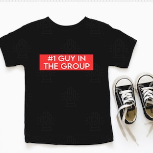 May include: Black t-shirt with a red rectangle that says "#1 GUY IN THE GROUP" on it. The shirt has a small white hand print design repeated all over it.