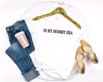 May include: White t-shirt with the text "IN MY MOMMY ERA" printed in black on the front. The shirt is on a white fluffy surface with a gold sequin hanger, a pair of blue jeans, and a pink coffee cup.