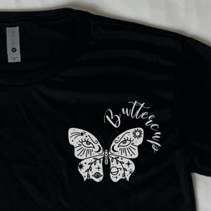 May include: Black t-shirt with a white butterfly graphic and the word "Butterfly" written in cursive. The butterfly has celestial details like stars, moons, and eyes.