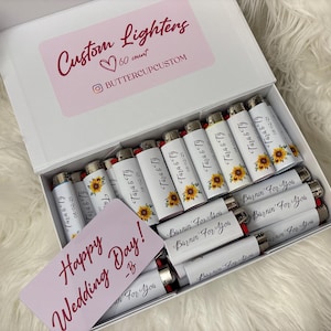 May include: A white box filled with custom lighters, each with a unique design. Some lighters feature sunflower graphics and the names "Tara & Ty" with a date. A card reads "Happy Wedding Day!" and "Burnin' For You."