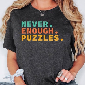 May include: A dark gray t-shirt with the text "NEVER. ENOUGH. PUZZLES." printed in colorful letters. The text is in a retro style font.