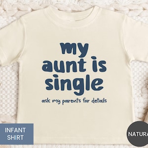 May include: Cream-colored infant shirt with the navy blue text "my aunt is single" and "ask my parents for details." The shirt has short sleeves and a crew neck. The words "Infant Shirt" are in a blue box, and "Natural" is in a black circle.