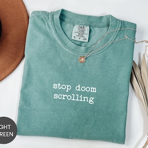 May include: A light green t-shirt with the text "stop doom scrolling" in white. The shirt has a crew neck and short sleeves. A gold necklace with a pendant rests on the shirt. The shirt is folded on a white surface.