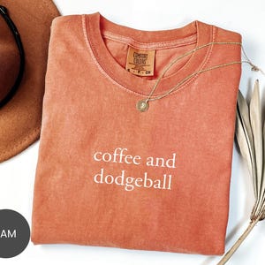 May include: A coral-colored t-shirt with the words "coffee and dodgeball" in white script. A gold necklace with a pendant rests on the shirt. A brown hat and dried palm leaves are in the background.