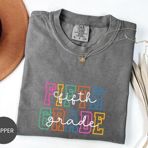 May include: A charcoal gray short-sleeve t-shirt with a crew neckline. The front features the text FIFTH GRADE in colorful block letters with the words fifth grade written in white cursive script layered over the top. A small tag is visible at the neck.