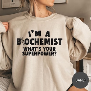 May include: Sand-colored sweatshirt with the text "I'M A BIOCHEMIST WHAT'S YOUR SUPERPOWER?" in black. The word "BIOCHEMIST" has a DNA strand graphic. The sweatshirt has a crew neck and long sleeves.