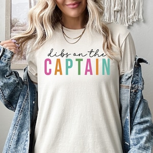 May include: A beige t-shirt with the text "dibs on the CAPTAIN" in colorful letters. The letters are pink, orange, green, and teal.