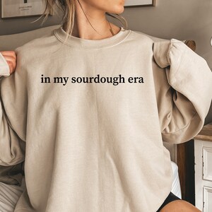 May include: A beige sweatshirt with the text "in my sourdough era" printed in black.