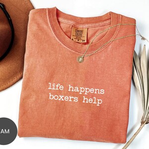 May include: A coral-colored t-shirt with the phrase "life happens boxers help" in white lettering. A gold necklace with a pendant rests on the shirt. A brown hat is visible in the upper left corner.