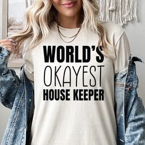 May include: Beige t-shirt with the text "WORLD'S OKAYEST HOUSE KEEPER" in bold black letters. The person is wearing a denim jacket over the shirt. The shirt is a casual wear item.