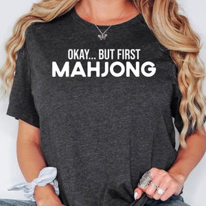 May include: Dark gray t-shirt with the text "Okay... But First Mahjong" in white block letters. The shirt is a crew neck style and appears to be made of a soft material. The overall theme is related to games and leisure.