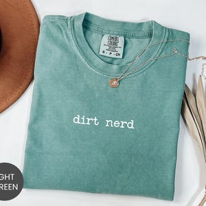 May include: Light green t-shirt with the words "dirt nerd" printed in white. The shirt has a crew neck and short sleeves. A silver necklace with a round pendant is draped across the shirt.