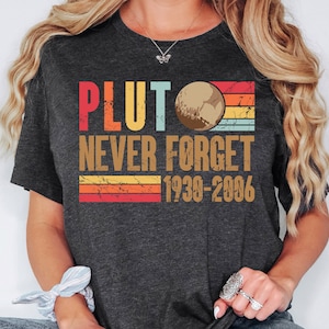 May include: A dark grey t-shirt with a vintage-style graphic that says "Pluto Never Forget 1930-2006". The graphic features a stylized image of Pluto with a rainbow stripe background.